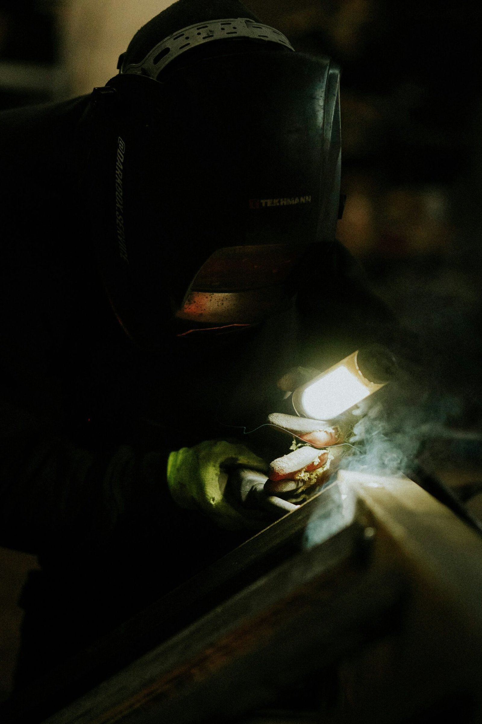 Close-up of a welder in a dark workshop using a flashlight and precision tools for detailed welding, with smoke and glow highlighting the scene.
