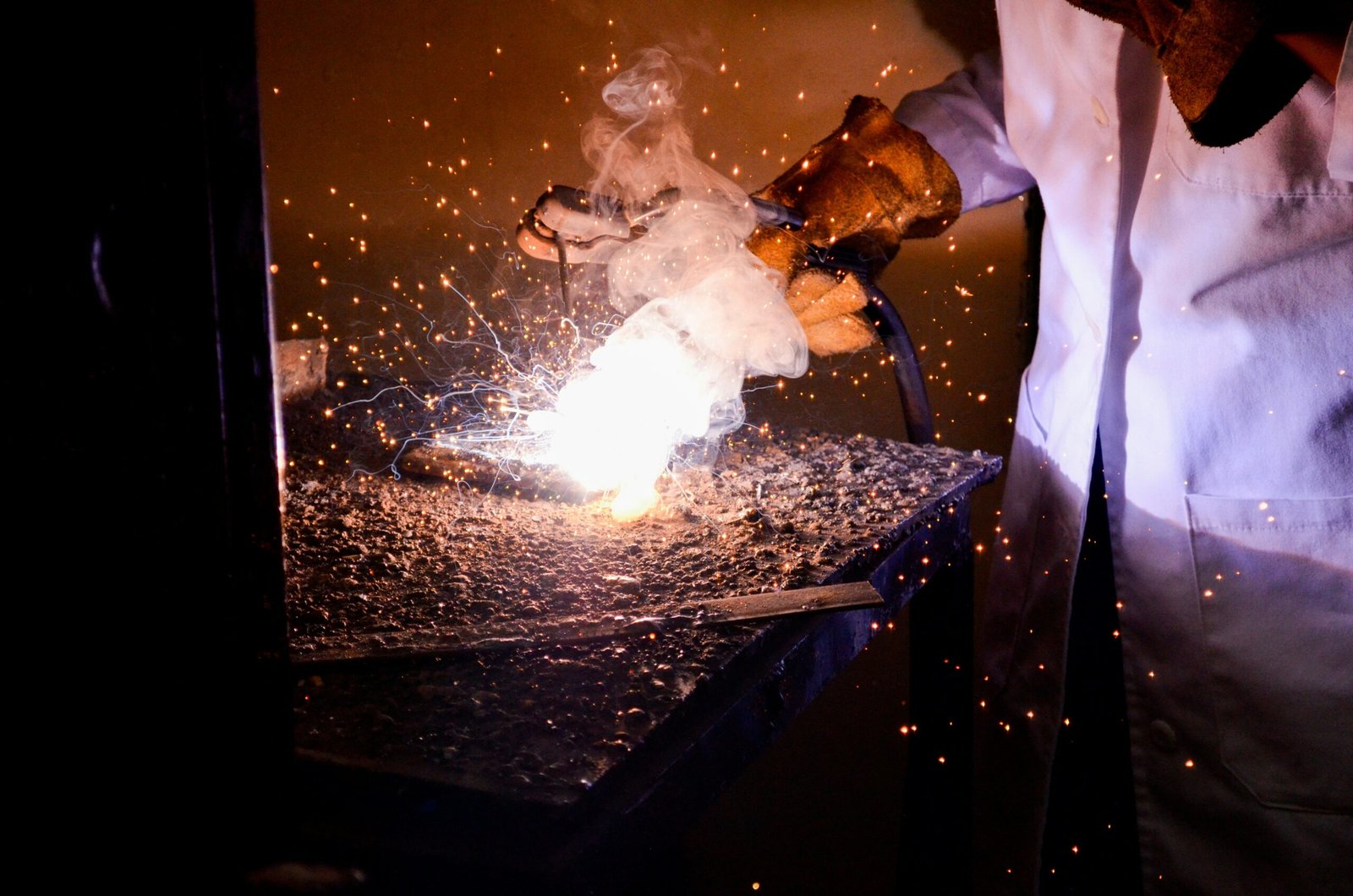 Close-up of a welder in a white lab coat and leather gloves performing arc welding on a metal surface, with intense sparks and smoke rising.
